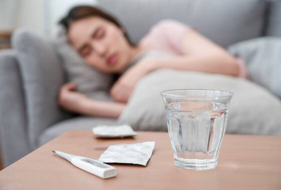 Zdjęcie kobiety, którą rozłożyło przeziębienie. Chora leży w łóżku, przed nią na pierwszym planie szklanka wody i termometr. Zdjęcie przykładowe.