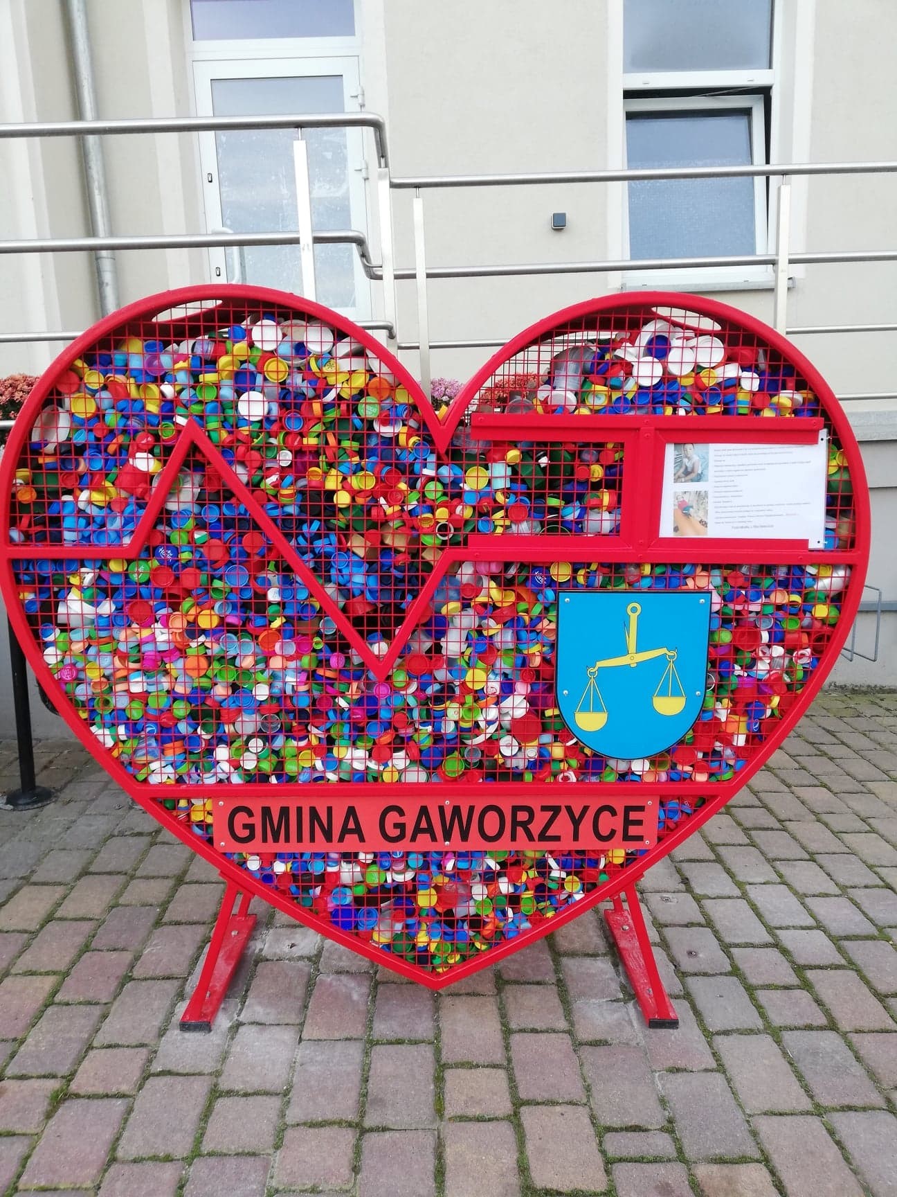 Zdjęcie kosza w kształcie serca na nakrętki przed budynkiem GOPS w Gaworzycach
